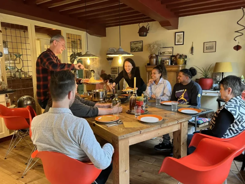 Diverse groep mensen die lachend samen spaghetti eten aan een grote houten tafel met rode stoelen, in een warm, huiselijk interieur — verbinding en ontmoeting bij de Red Chair Movement.