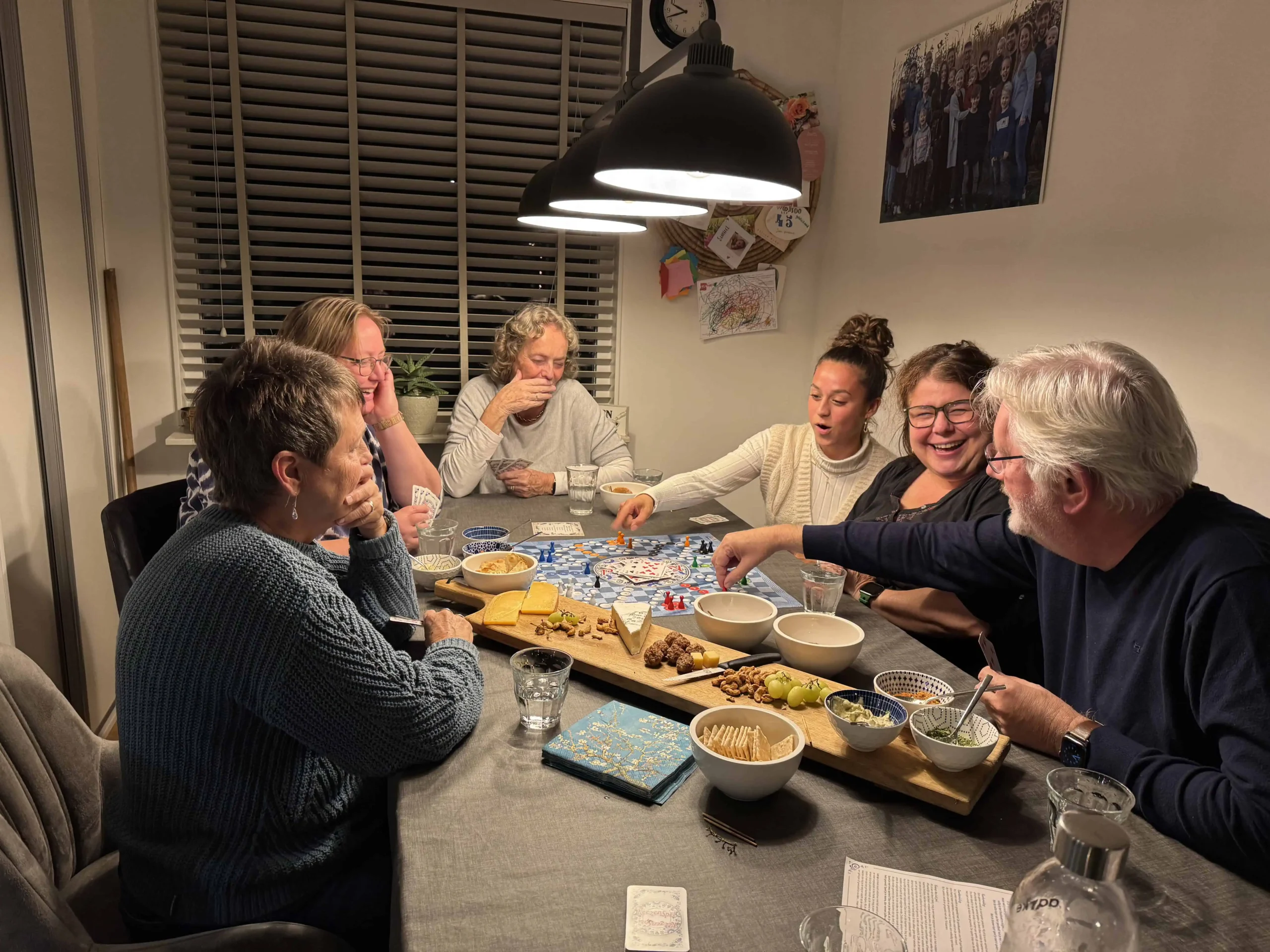 Groep mensen die lachend samen een bordspel spelen aan een gezellig gedekte tafel met borrelplank, tijdens een warme spelletjesavond — verbinding en ontmoeting bij de Red Chair Movement.